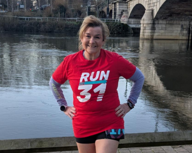 Women in red t shirt bearing the logo Run 31 with hands on hips, facing camera. There is a river behind her